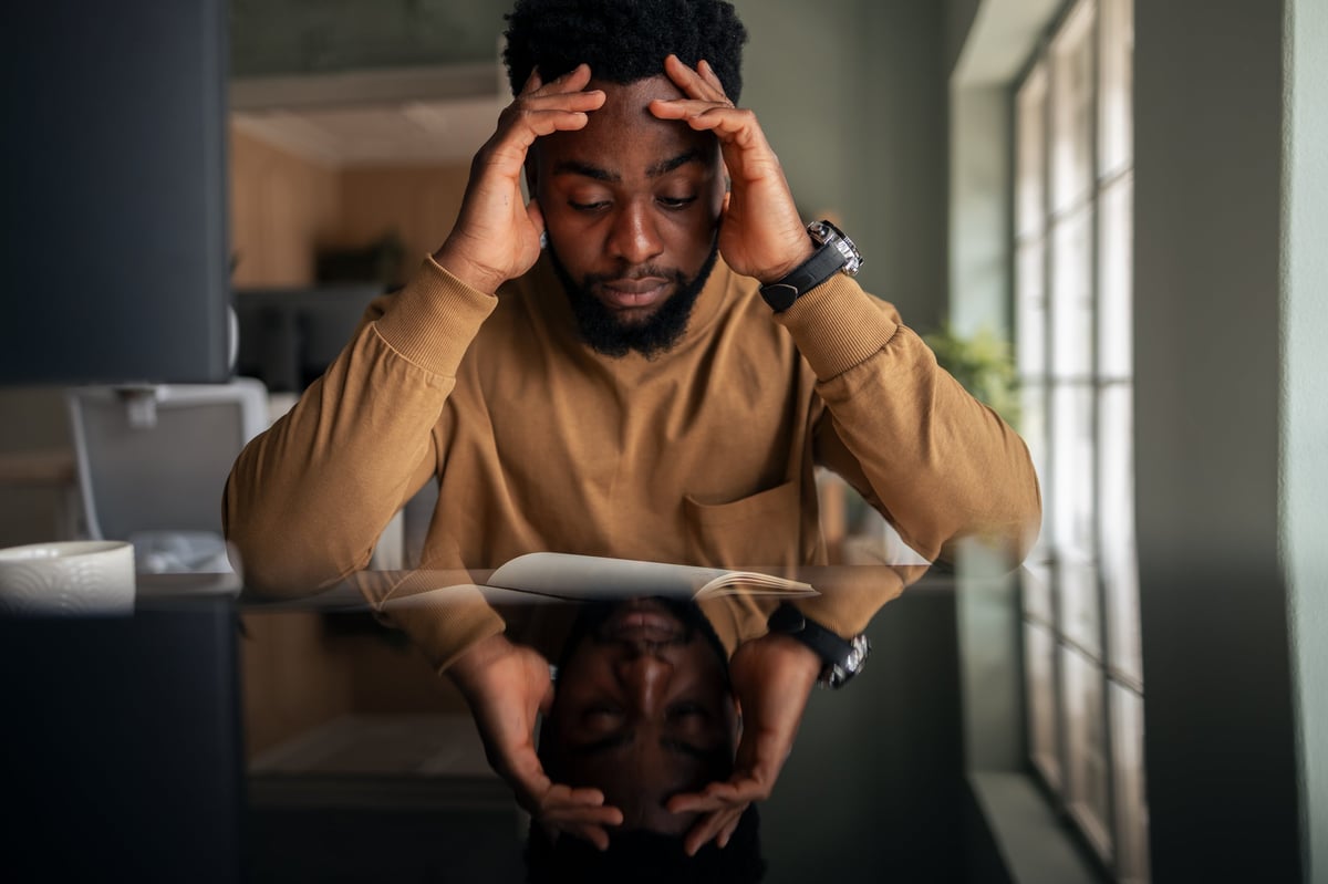 A contemplative individual sits at a desk, brows furrowed, deeply lost in thought, his reflection visible on the desk's surface, evoking focus and introspection in a modern office space.