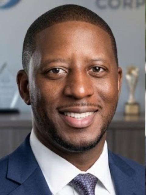Professional headshot of a man in a dark blue suit and tie, smiling at the camera against a blurred office background.